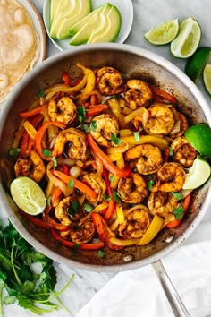 a skillet filled with shrimp and peppers next to tortillas, avocado wedges and cilantro