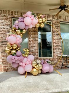 a balloon arch with pink and gold balloons in front of a brick building that has a large number on it