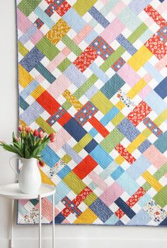 a colorful quilt hanging on the wall next to a white chair and vase with flowers