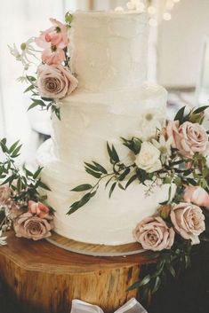 a white wedding cake with pink flowers and greenery on the top is sitting on a wooden stand