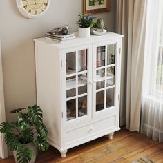 a white cabinet sitting next to a window with a clock on the wall above it