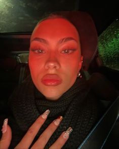 a woman with bright red makeup and nails in a car looking surprised at the camera