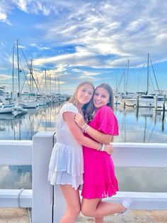 two girls hugging each other in front of a marina with boats on the water behind them