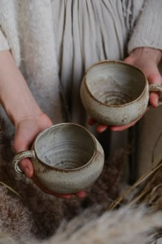 a person holding two mugs in their hands with dried grass behind them and brown fur on the floor