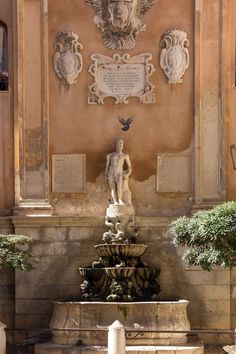 a fountain in front of a building with statues on the wall and trees around it