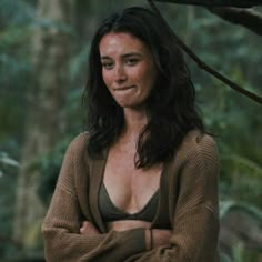 a woman standing in the woods with her arms crossed and looking at the camera while wearing a brown sweater