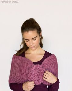 a woman wearing a purple sweater with a flower on the front and shoulder, standing in front of a white wall