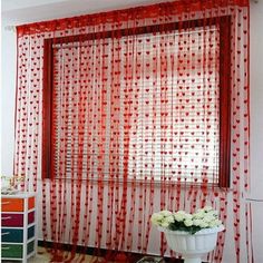 a vase with flowers sitting in front of a window covered in red sheer curtaining