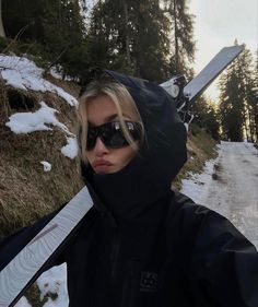 a woman in sunglasses holding a snowboard on the side of a snowy mountain road