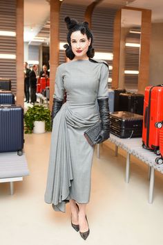 a woman in a gray dress and black gloves stands next to luggage at an airport