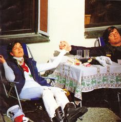 two men sitting at a table with food and drinks in front of them on folding chairs