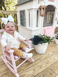 a baby sitting in a pink rocking chair on a wooden deck next to a doll house