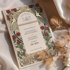 an ornate wedding card with flowers and leaves on white satin, surrounded by dried grasses