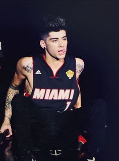 a man with tattoos sitting on top of a basketball court wearing a black and red uniform