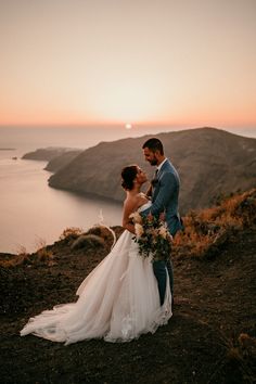 a bride and groom standing on top of a hill next to the ocean at sunset