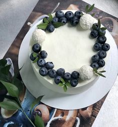 a white cake with blueberries and green leaves on it sitting on top of a magazine