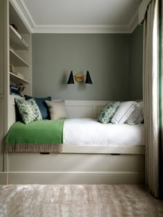 a bed sitting under a window next to a book shelf filled with books and pillows