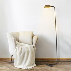 a white chair with a blanket on it next to a floor lamp and wooden floor