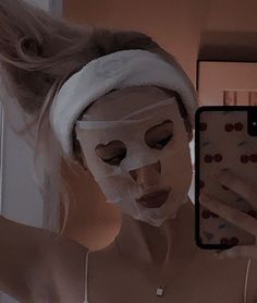 a woman wearing a white mask looking at her cell phone in front of the mirror