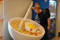 a man standing next to a giant bowl of noodles with an egg in it