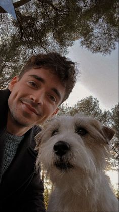 a man sitting next to a white dog on top of a forest covered ground with trees in the background