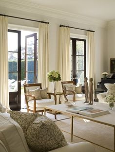 a living room filled with lots of furniture next to two large windows covered in white curtains