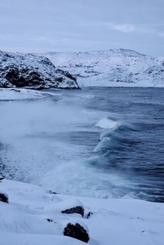 a body of water with snow on the ground and mountains in the background, near some rocks