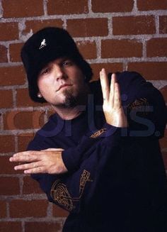 a man wearing a black hat and holding his hands up in front of a brick wall