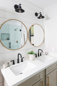 two round mirrors hanging on the wall in a white bathroom with black faucets