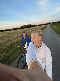 two women riding bicycles on a road with grass and trees in the background at sunset