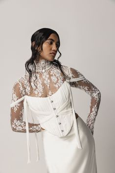 a woman wearing a white dress with sheer sleeves and lace on the top, standing in front of a gray background