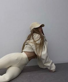 a woman sitting on the ground wearing white pants and a hat with her hair blowing in the wind