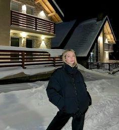 a woman standing in front of a house covered in snow at night with the lights on