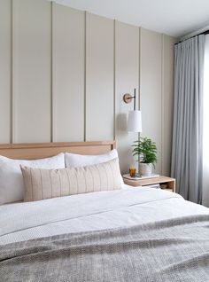 a bed with white sheets and pillows next to a lamp on a nightstand in front of a window