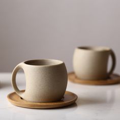 two coffee cups sitting next to each other on top of a white table with wooden coasters