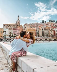 a man and woman kissing on the edge of a bridge over water with buildings in the background