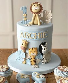 a blue cake with animals and letters on it sitting on a table next to cupcakes