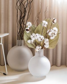 two white vases with flowers in them on a table next to a curtained window