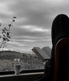 a person sitting in a chair with a book and cup on the window sill