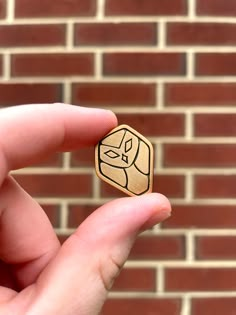 a hand holding a small wooden object with a cat on it's face in front of a brick wall