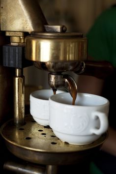 two coffee cups being filled with liquid from an espresso machine on a gold plate