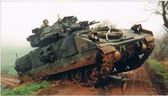 two men riding on top of an army tank down a dirt road in the woods