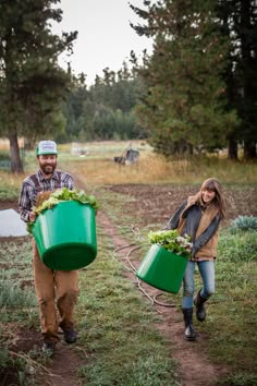 two people carrying green buckets full of lettuce in a field with trees