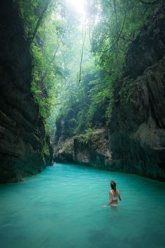 a woman standing in the middle of a river surrounded by lush green trees and rocks