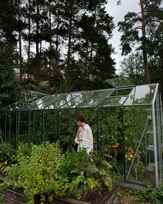 a woman standing in front of a green house surrounded by plants and trees, looking down at the ground