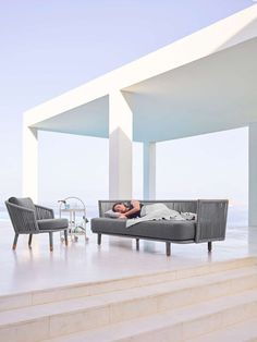 a man laying on top of a gray couch next to a white building near the ocean