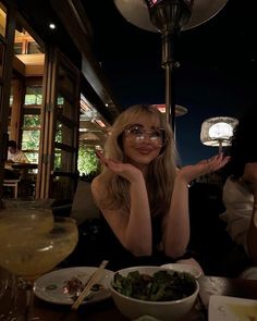 a woman sitting at a table in front of a bowl of food with her hands up