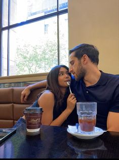 a man and woman sitting at a table with drinks in front of them, one is biting into the other's mouth