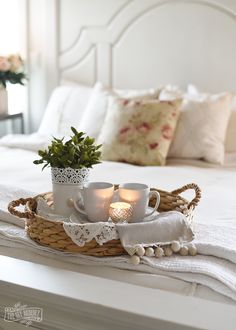 two cups on a tray with tea lights in them sitting on a bed that has white linens and pillows