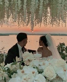 a bride and groom sitting at a table with flowers in front of the sun setting